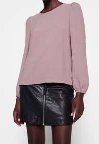 Blouse à manches longues couleur mauve clair avec tissu texturé et coutures d'épaules froncées, associée à une mini-jupe en cuir noir dotée d'une fermeture éclair latérale.