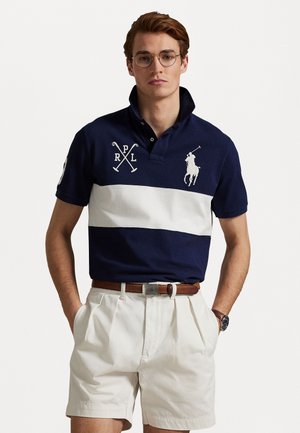 Navyblaues Poloshirt mit weißen horizontalen Streifen, gesticktem Logo und überkreuzten Schlägern. Kombiniert mit beigen Falten-Shorts und einem braunen Gürtel.