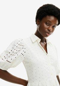 Abito bianco in cotone con maniche corte a sbuffo, motivo ad occhio di pernice, colletto e chiusura con bottoni, caratterizzato da una gonna fluida e dettagli testurizzati.