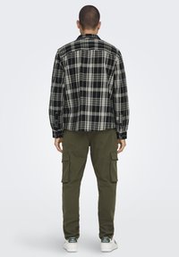 Cămașă din flanel în carouri alb-negru, cu mâneci lungi, asociată cu pantaloni cargo verzi măslinii și adidași albi cu accente verzi.