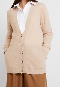 Cárdigan beige con escote en V, mangas largas y botones en la parte delantera. Tiene dos bolsillos frontales y una tela suave y texturizada.