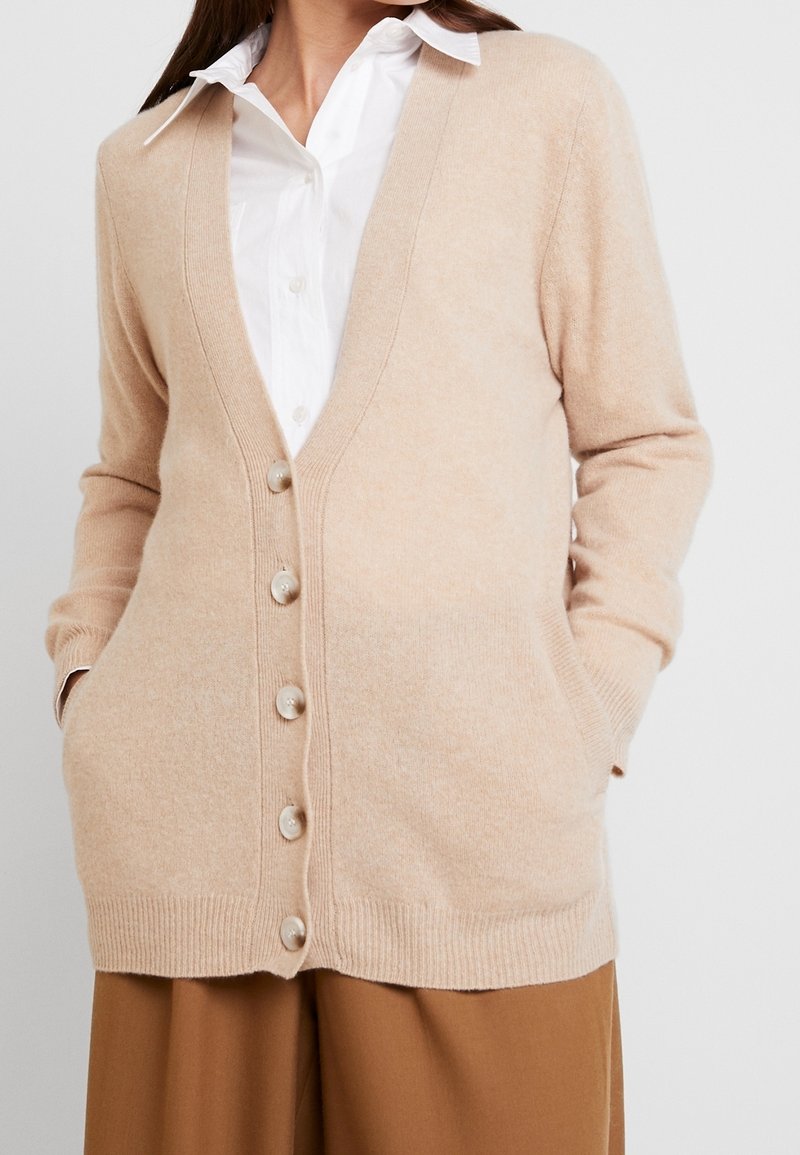 Cárdigan beige con escote en V, mangas largas y botones en la parte delantera. Tiene dos bolsillos frontales y una tela suave y texturizada.