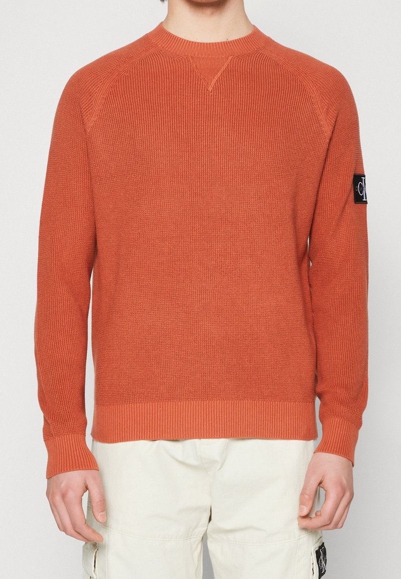 Orange stickad tröja med ribbad halsringning och ärmslut, med en svart logotypapplikation på vänstra ärmen. Slät textur, figurnära design.