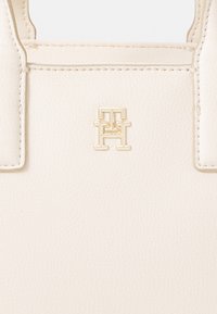Borsa in pelle color crema con superficie texturizzata, logo del brand in tonalità oro e manici superiori rinforzati. Design minimalista con linee pulite.