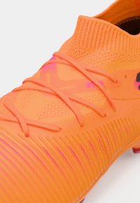 Jaskrawo pomarańczowy but piłkarski z teksturowanym syntetycznym wierzchem, misternym wzorem sznurowadeł i żebrowanym wzorem po bokach, z różowymi akcentami.