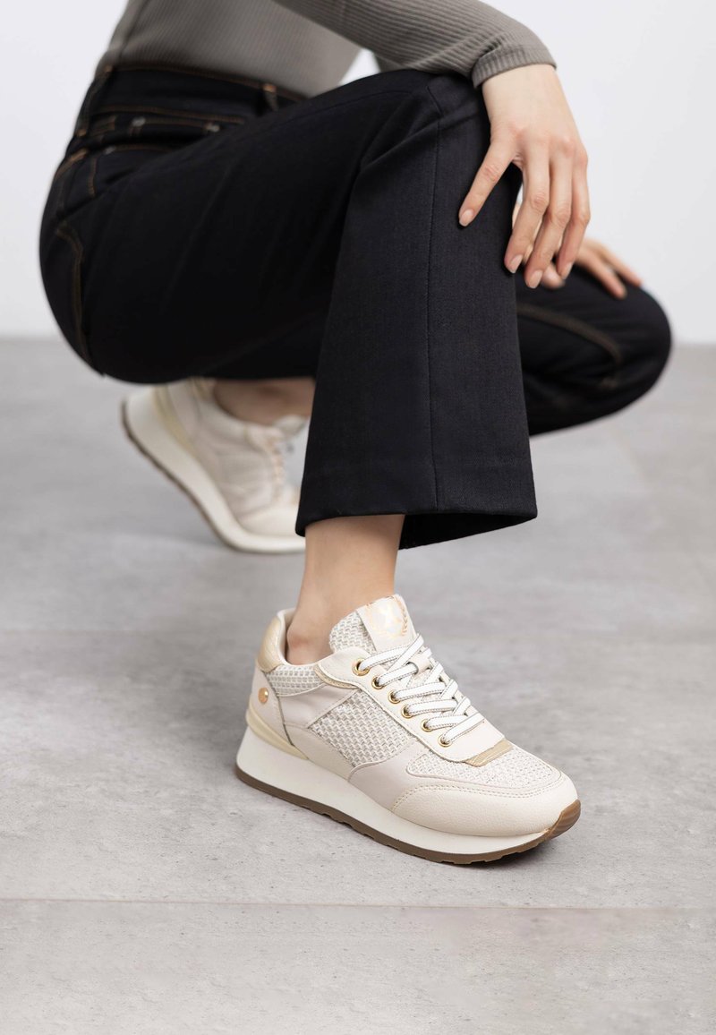 Zapatillas beige claro con paneles de malla y cuero, punta redonda y cordones blancos. Detalles texturizados y suela de goma en marrón contrasta.