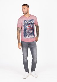 Rode T-shirt met korte mouwen met een motorfietsafbeelding, gecombineerd met grijze distressed slim-fit jeans en witte sneakers.
