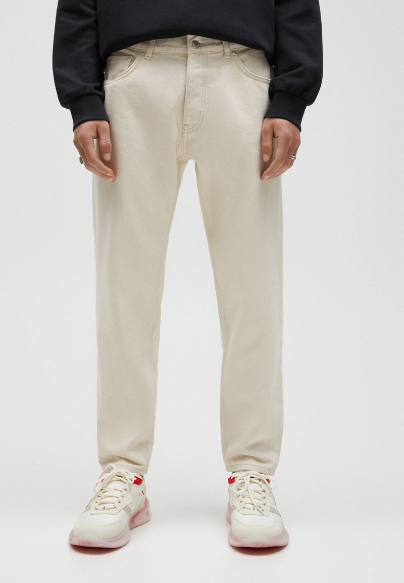 Béžové džíny s rovnými nohavicemi, pětikapsovým designem a prošitými detaily, kombinované s bílými teniskami s růžovými akcenty.