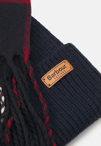 Tengerészkék sál bordás mintával, piros és fekete kockás díszítéssel, valamint egy téglalap alakú barna bőrcímkével, amelyen a "Barbour England" felirat látható.