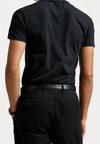 Maglietta nera in cotone con maniche corte, caratterizzata da un design aderente e una texture liscia, abbinata a pantaloni scuri e una cintura in pelle nera.