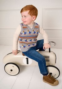 Niño con un chaleco de punto gris con un patrón de patos sentado en un coche de juguete beige con ruedas negras, con el número blanco "1" en el lateral, vistiendo unos vaqueros azules y zapatos marrones.