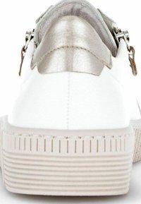 Baskets blanches avec un accent métallique argenté au talon, semelle texturée et fermeture à glissière sur le côté. Forme arrondie et finition lisse.