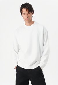 Sweatshirt branco com gola redonda, punhos e cós canelados. Tecido suave, ajustamento descontraído, design simples, sem logótipos ou padrões visíveis.