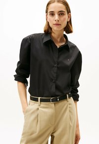 Zwarte button-up shirt met opgerolde mouwen, voorzien van een klein logo, gecombineerd met beige op maat gemaakte broeken en een zwarte riem met een zilveren gesp.
