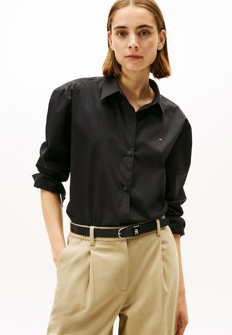 Zwarte button-up shirt met opgerolde mouwen, voorzien van een klein logo, gecombineerd met beige op maat gemaakte broeken en een zwarte riem met een zilveren gesp.