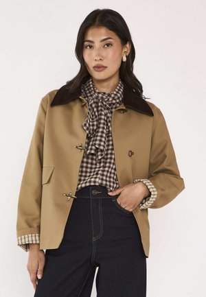 Woman wearing a tan jacket over a brown checkered blouse with a bow, paired with dark jeans, posing with one hand in pocket.