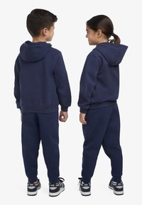 Conjunto de moletom azul-marinho e calças de fato feito de material suave. Design com capuz e punhos canelados. Usado com sapatilhas pretas e brancas.