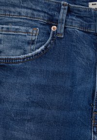 Jean en denim de couleur bleu délavé, avec une poche avant, un rivet en cuivre et une texture de couture visible. Ourlet usé à la taille.
