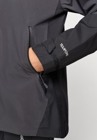 Veste imperméable noire avec une poche zippée sur la manche et une bande Velcro, tissu texturé à finition lisse, marquage réfléchissant.
