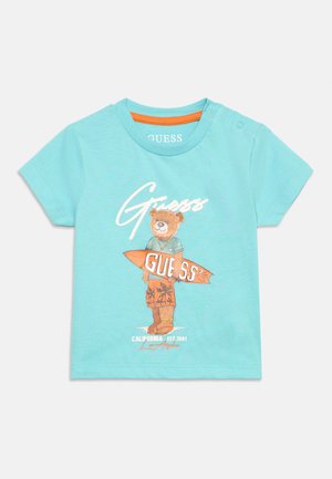 T-shirt bleu clair pour tout-petit avec un ours tenant une planche de surf orange marquée "Guess" et le texte "California Est. 1981 Los Angeles."