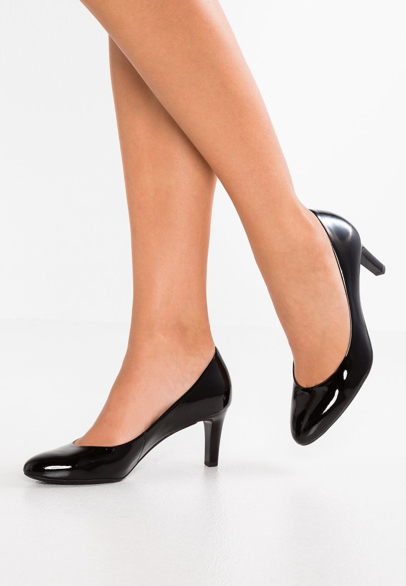 Jambes portant des escarpins noirs brillants à talons moyens, un pied à plat sur le sol, l'autre légèrement relevé sur un fond blanc.