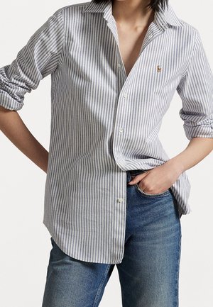 Personne portant une chemise boutonnée à rayures verticales bleues et blanches avec manches retroussées et un jean bleu, une main dans la poche.