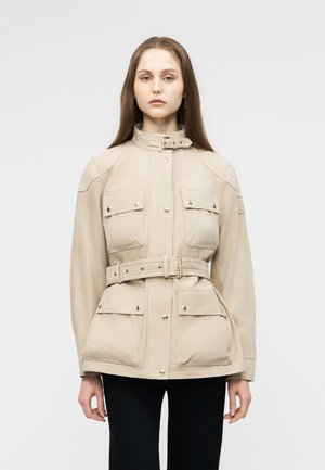 Žena s dlhými priamymi vlasmi oblečená v béžovom páse s štyrmi vreckami s chlopňami a čiernych nohaviciach na jednoliatom pozadí.