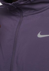 Nahaufnahme einer violetten perforierten Zip-Jacke mit Kapuze und weißem Nike-Swoosh-Logo auf der Brust.