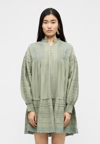 Νεαρή γυναίκα με ίσια σκούρα μαλλιά, φοράει ένα μακρυμάνικο φόρεμα σε χρώμα φασκόμηλο με περίπλοκα δαντελένια και πιετέ στοιχεία, στέκεται μπροστά σε απλό φόντο.