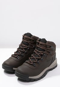 Botas de senderismo de cuero marrón con acentos de malla negra, que cuentan con ojetes reforzados, cordones marrones y una suela de goma texturizada. Diseñadas para uso al aire libre.