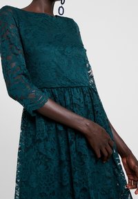 Tee-Farbenes Spitzenkleid mit langen durchsichtigen Ärmeln, floralem Muster, geraffter Taille und schwingendem Rock. Detail des Ärmels und der Hand sichtbar.