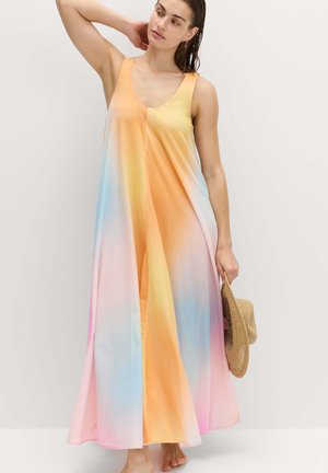 Kvinde iført en ærmeløs, flydende maxi-kjole med en pastelgradient i orange, gul, blå og pink, der holder en stråhat i den ene hånd.