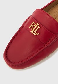 Rdeč usnjen loafer z všitimi detajli in zlatim logotipom. Gladek tekstura in zaobljena oblika, primerna za priložnostno nošenje.