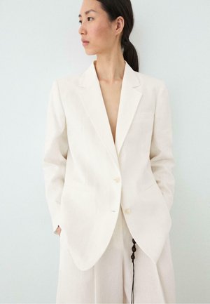 Kvinde med mørkt hår bundet bagud iført en løs hvid blazer og matchende vidde bukser, ser til venstre med hænderne i lommerne.