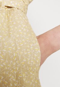 Gul blommönstrad tyg med vita och ljusgula blommor på en beige bakgrund, med en slät textur och avslappnad passform.