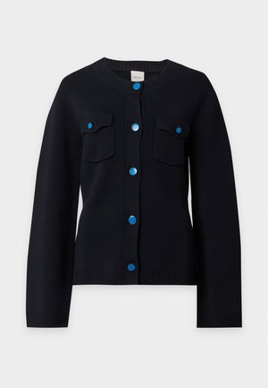 Persoon die een zwarte cardigan met blauwe knopen draagt, grijze wijde broek en zwarte puntige schoenen, staand met handen in de zakken tegen een effen achtergrond.