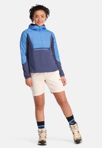 Blau und marineblaues Kapuzenjackett mit einem halben Reißverschluss, kombiniert mit hellen Shorts und braunen Wanderschuhen, akzentuiert durch dunkelblaue und weiße gestreifte Socken.