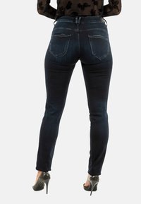 Jeans de mezclilla oscuros y ajustados con una cintura de media altura, que cuentan con dos bolsillos traseros y detalles de costura sutiles. Llevados con zapatos de tacón alto.