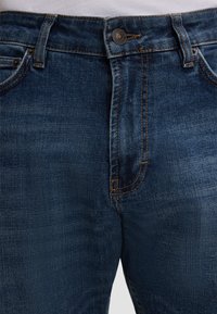 Granatowe dżinsy o gładkiej fakturze, z zapięciem na guzik, metalowymi detalami i kontrastowymi pomarańczowymi przeszyciami wzdłuż szwów.