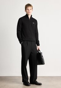 Sweater preto com fecho, com um pequeno logótipo, combinado com calças pretas de pregas e sapatos de couro pretos, segurando uma mala preta.