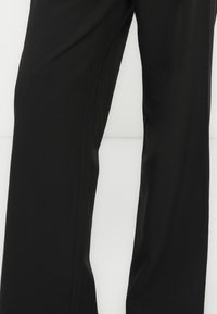 Gros plan d’un pantalon noir à coupe droite montrant la texture du tissu et les coutures latérales sur un fond clair uni.