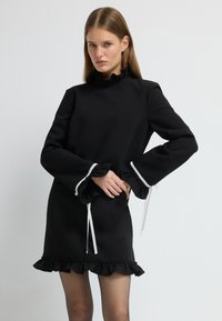 Černé šaty s dlouhým rukávem, zdobené volánkovým límečkem a lemem. Má bílé šňůrky na rukávech. Hladký materiál a přiléhavý střih.