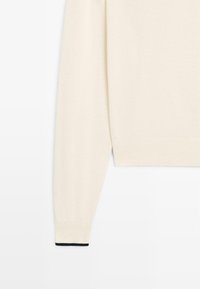 Pull à manches longues couleur crème avec poignets et ourlet côtelés, présentant un petit liseré noir au niveau du poignet.