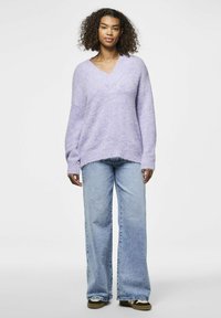 Liliowy, oversize sweter z dekoltem w kształcie V, teksturowana dzianina, w zestawieniu z jasnoniebieskimi spodniami z szerokimi nogawkami i ciemnymi butami.