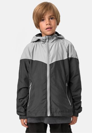 Junge mit hellbraunen Haaren, der eine graue und schwarze Kapuzenjacke mit Reißverschluss trägt, steht vor einem schlichten weißen Hintergrund.