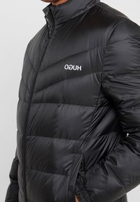 Sort puffet jakke med horisontalt quiltet mønster, høj krave, frontlynlås og hvid "HUGO" logo på venstre bryst. Glat, syntetisk materiale.