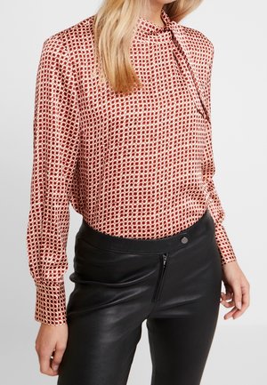 Mujer con blusa de manga larga, con estampado rojo y blanco y detalle de cuello anudado, combinada con pantalones negros de cuero de tiro alto.