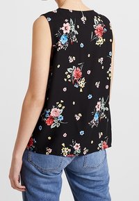 Ärmelloses schwarzes Oberteil mit buntem Blumenmuster, getragen mit blauen Jeans, von hinten gezeigt auf einer Person, die vor einem schlichten Hintergrund steht.