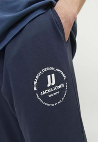Mörkblå mjukisbyxor med en vit cirkulär logotyp som visar "JACK & JONES" och text, tillverkade av ett mjukt, strukturerat tyg. Fickan synlig.