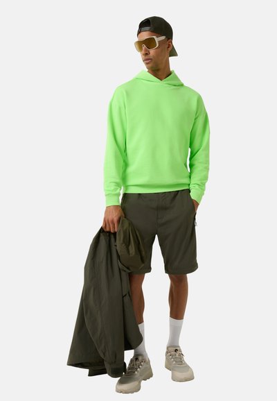 Neon zielona bluza, oliwkowe szorty i jasnoszare sneakersy. Trzyma oliwkową kurtkę. Sportowe okulary przeciwsłoneczne, krótki rękawek.